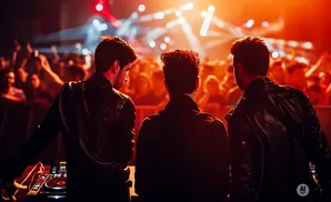 Three DJs stand facing a cheering crowd under bright, red stage lights at a concert.