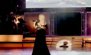 Singer on stage holding microphone, wearing a black dress with a sparkly overlay top.