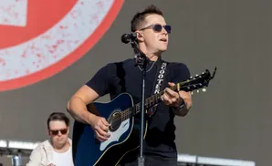 A man with sunglasses plays an acoustic guitar on stage, with another man blurred in the background.