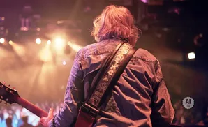 Guitarist in denim shirt on stage with warm lighting and audience blurred in the background.