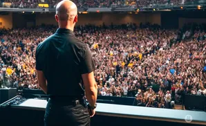 A bald man in a black shirt stands on stage facing a large, cheering crowd.