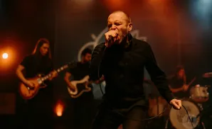 A male singer in a black shirt sings into a microphone on stage, with bandmates playing guitar and drums behind him.