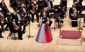 A conductor in a colorful skirt stands with a violin in front of an orchestra.