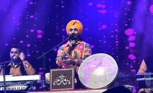 Musicians performing on stage with keyboard, drum, and traditional Indian instrument, under purple stage lighting.