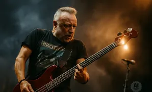 A male bassist with a gray beard plays a red electric bass guitar on stage under warm lighting.