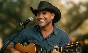A smiling man in a cowboy hat plays an acoustic guitar and sings into a microphone outdoors.