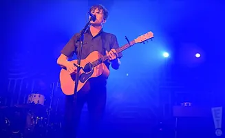A man with dark hair wearing a dark shirt plays an acoustic guitar and sings into a microphone on a stage bathed in blue light.