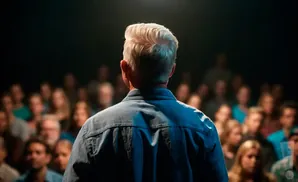 Man with slicked-back hair in a blue shirt addresses a blurred audience.