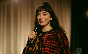 A woman with dark hair and a bun smiles widely while holding a microphone and wearing a red and black patterned shirt.
