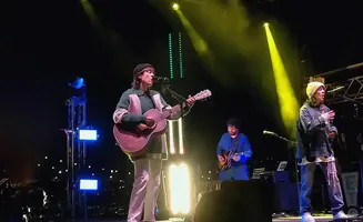 A band performs on stage at night, with bright spotlights and audience lights in the background.