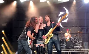 A rock band performs on stage with bright lights and a backdrop featuring a circular logo.