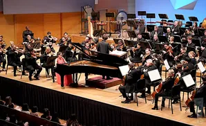 A pianist in a pink dress plays a grand piano with a symphony orchestra behind her.