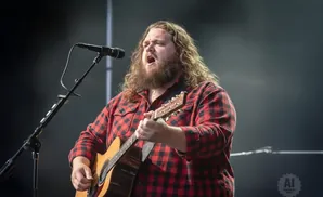 Singer with long, curly brown hair and a beard sings into a microphone while playing an acoustic guitar. He wears a red and black plaid shirt.