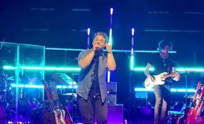 A man sings into a microphone on stage, while another plays a guitar.