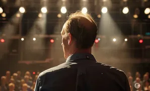 A man in a dark shirt stands on a stage, facing an audience, with spotlights and smoke behind him.