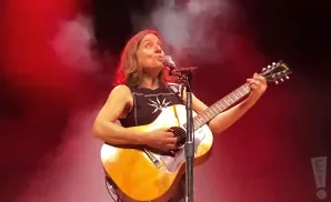 A woman plays an acoustic guitar on stage under red lights.