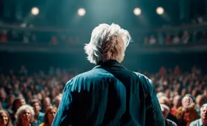 Person with white hair in a denim jacket stands on stage facing a large, dark audience.
