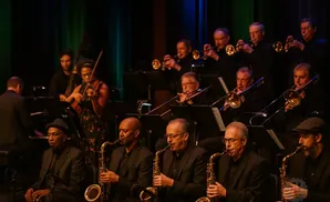 A jazz orchestra performs on stage, featuring saxophones, trombones, trumpets, and a violin.