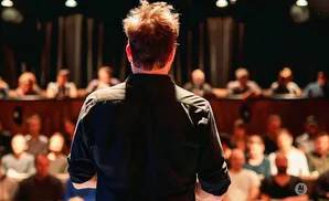Man with tousled hair on stage, back to the camera, facing a blurred audience in a dimly lit venue.
