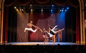 Dancers perform on a stage with red curtains, dramatic lighting, and an audience watching.
