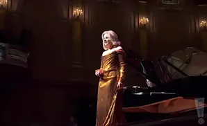 A woman in a golden gown sings on stage next to a grand piano.