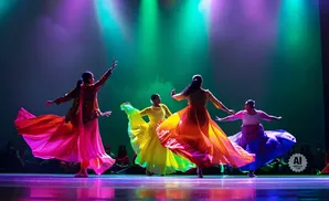 Dancers in colorful flowing dresses perform on a stage lit by purple and green lights.