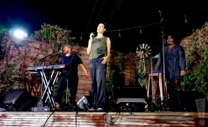 Three musicians perform on a stage at night, with a keyboardist on the left, a singer in the center, and a singer on the right.