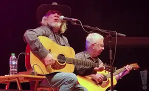 Musicians on stage playing acoustic guitars; one with a cowboy hat sings into a microphone.