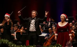 Conductor and singer in festive attire on stage with an orchestra during a holiday performance.