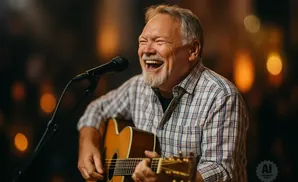 A man with a gray beard and plaid shirt laughs while playing an acoustic guitar and singing into a microphone.