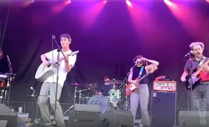 A band performs on stage with purple lighting. A man in a white t-shirt plays an acoustic guitar.