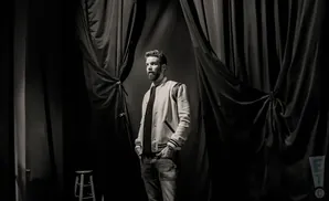 A man with a beard stands on a stage in front of dark curtains, wearing a letterman jacket.