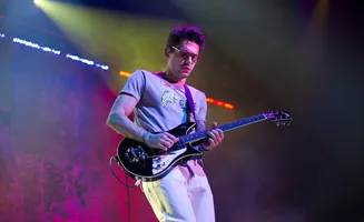 Musician playing a black guitar on stage with colorful lights.