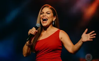 A woman in a red dress sings into a microphone with her mouth open and her right hand raised.