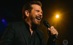 A man in a black suit sings into a microphone, laughing under a bright spotlight.