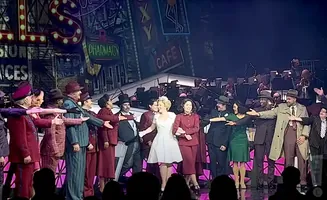 A cast of performers in vintage attire on stage, with a band in the background and a cityscape backdrop.