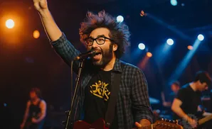 A man with wild hair and glasses sings into a microphone while playing guitar on stage.
