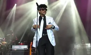 Performer in a white suit and tie sings into a microphone, with stage lights in the background.