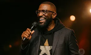 A Black man with a beard and glasses laughs while holding a microphone.