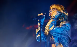 A woman sings into a microphone on stage, her eyes closed and hand to her cheek, illuminated by stage lights.