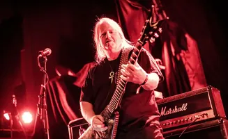 A guitarist with long white hair plays a dark electric guitar on a stage lit with red lights.