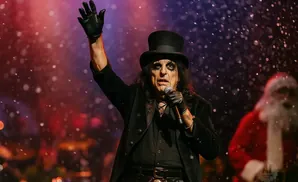 Alice Cooper in a top hat and black suit with theatrical makeup, raising his gloved hand on stage with Santa Claus blurred in the background.