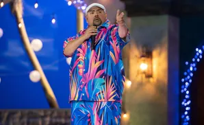 Comedian Gabriel Iglesias performs on stage wearing a colorful tropical shirt and pants.