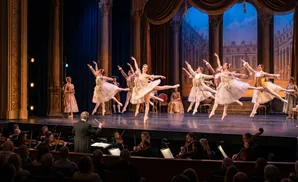 Ballet dancers in elaborate costumes perform on a stage with an orchestra in the foreground.