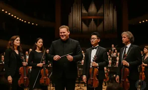 A conductor and violinists stand on stage before an orchestra, with a pipe organ in the background.