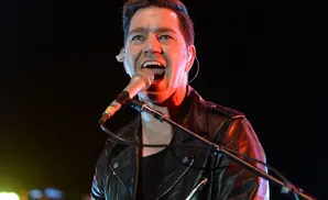 A man in a leather jacket sings into a microphone on a dark stage, lit by red and yellow lights.
