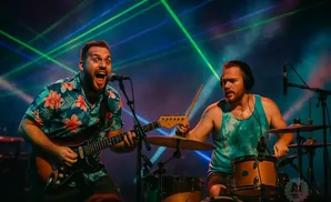 A band performs on stage with colorful lights shining from behind them. A singer plays guitar while a drummer plays behind him.