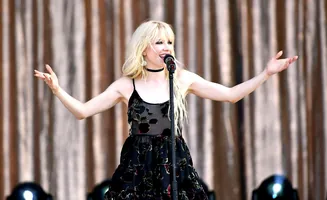 A blonde woman in a floral dress sings into a microphone on stage.