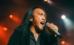 A male singer with long dark hair belts into a microphone, wearing a black velvet jacket and a cross necklace.