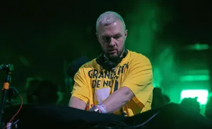 A DJ with blond hair and a yellow shirt plays music on a turntable under green lights.
